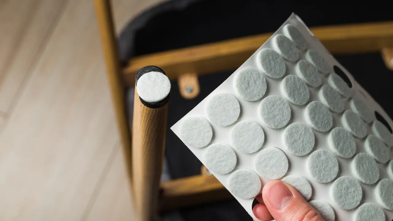 Close de uma mão aplicando um protetor de feltro adesivo no pé de uma cadeira de madeira para proteger o piso vinílico.