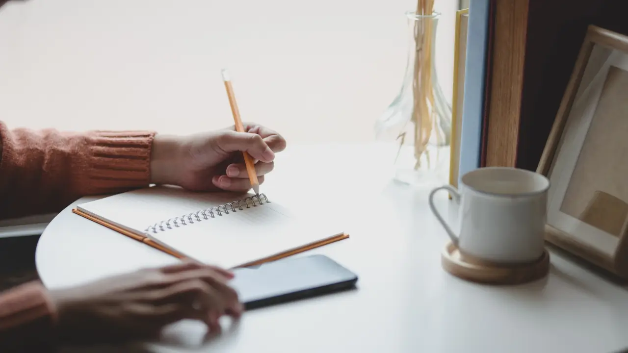 Mão escrevendo um plano de ação em um caderno ao lado de um smartphone com um app de organização aberto.