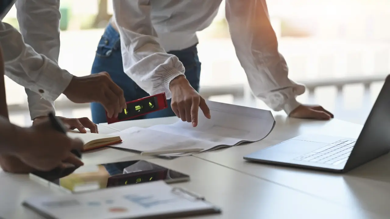 Mãos de um arquiteto sobre a planta de uma casa modular, com um computador portátil ao lado a mostrar o projeto em 3D.