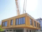 Processo de montagem de casas modulares, com uma grua a posicionar um módulo de uma habitação contemporânea num terreno arborizado.