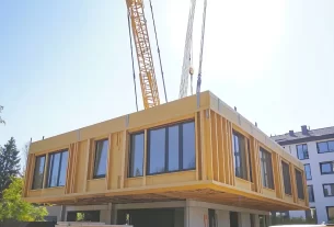 Processo de montagem de casas modulares, com uma grua a posicionar um módulo de uma habitação contemporânea num terreno arborizado.