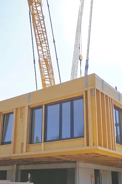 Processo de montagem de casas modulares, com uma grua a posicionar um módulo de uma habitação contemporânea num terreno arborizado.